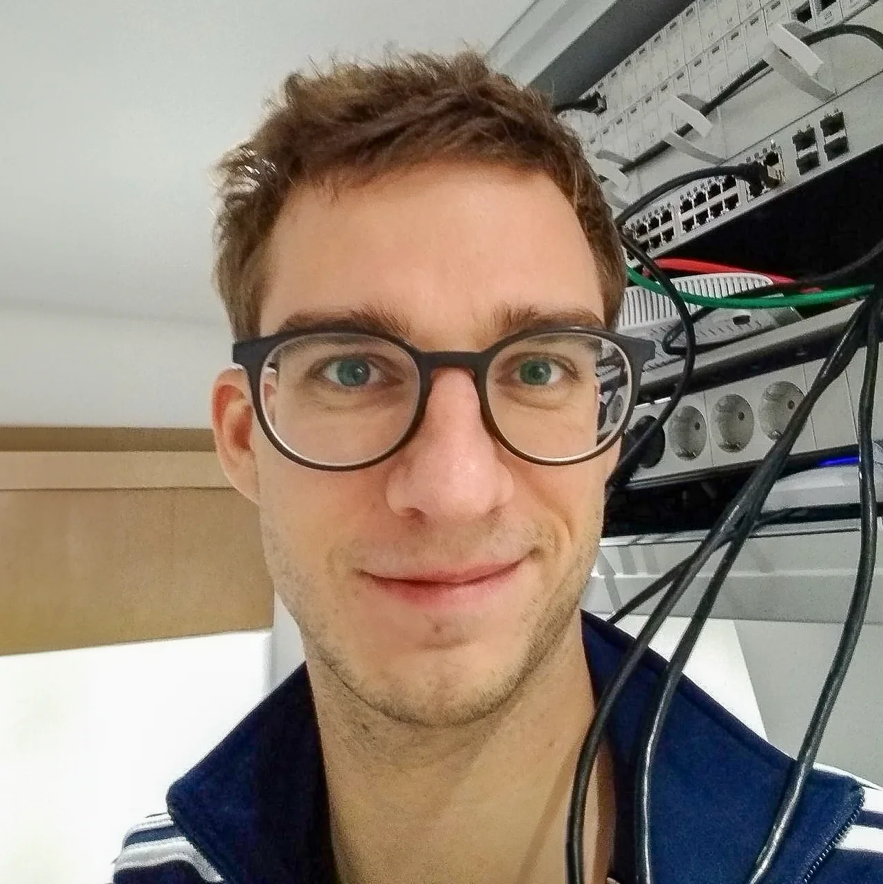 Foto von Eddie Görg vor einem Serverschrank. Netzwerkkabel kommen aus dem Serverschrank